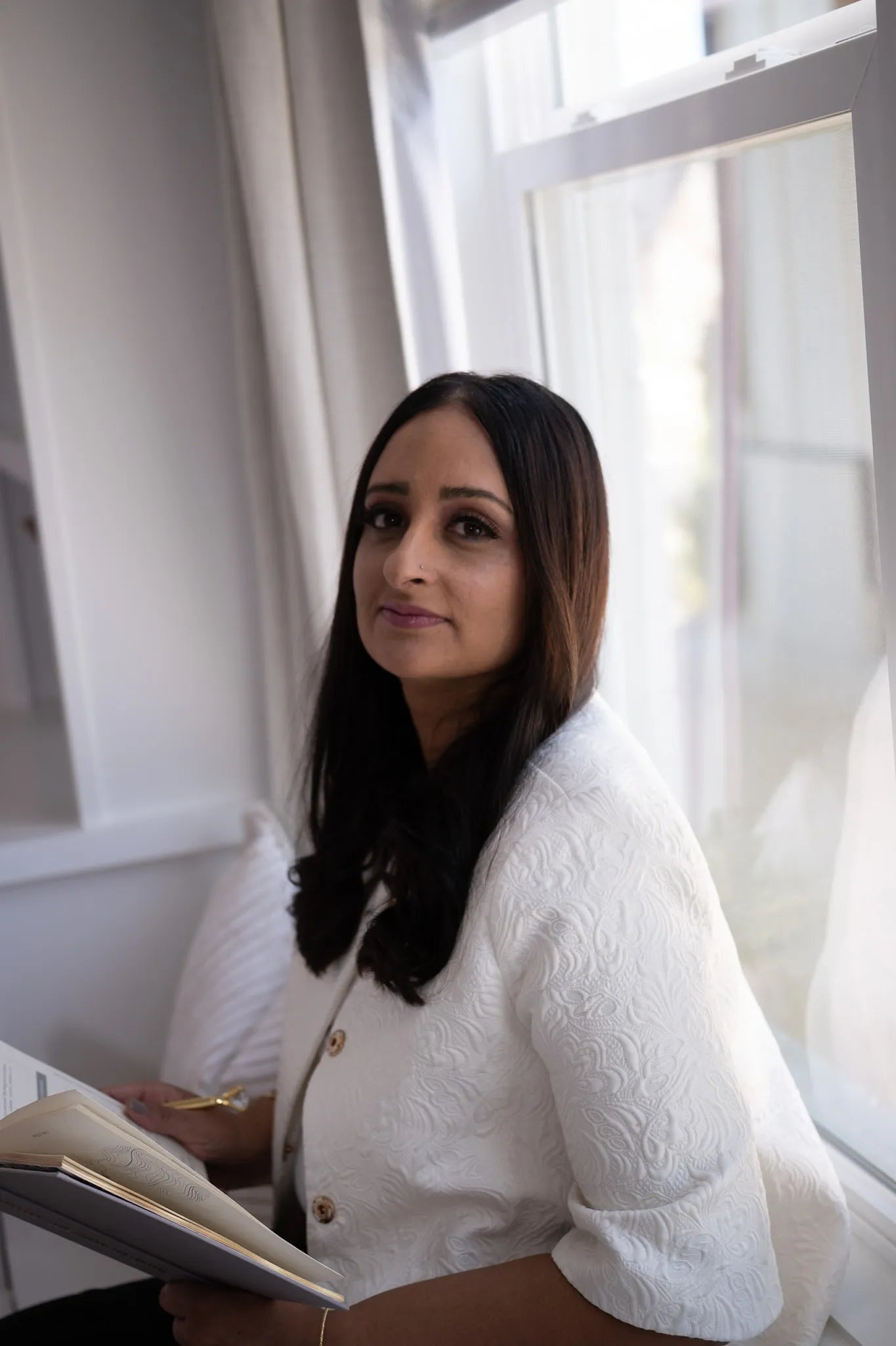Woman in a white textured jacket holding a book titled '.1 Daily Gratitude Journal for Lawyers' in a bright room with large windows.