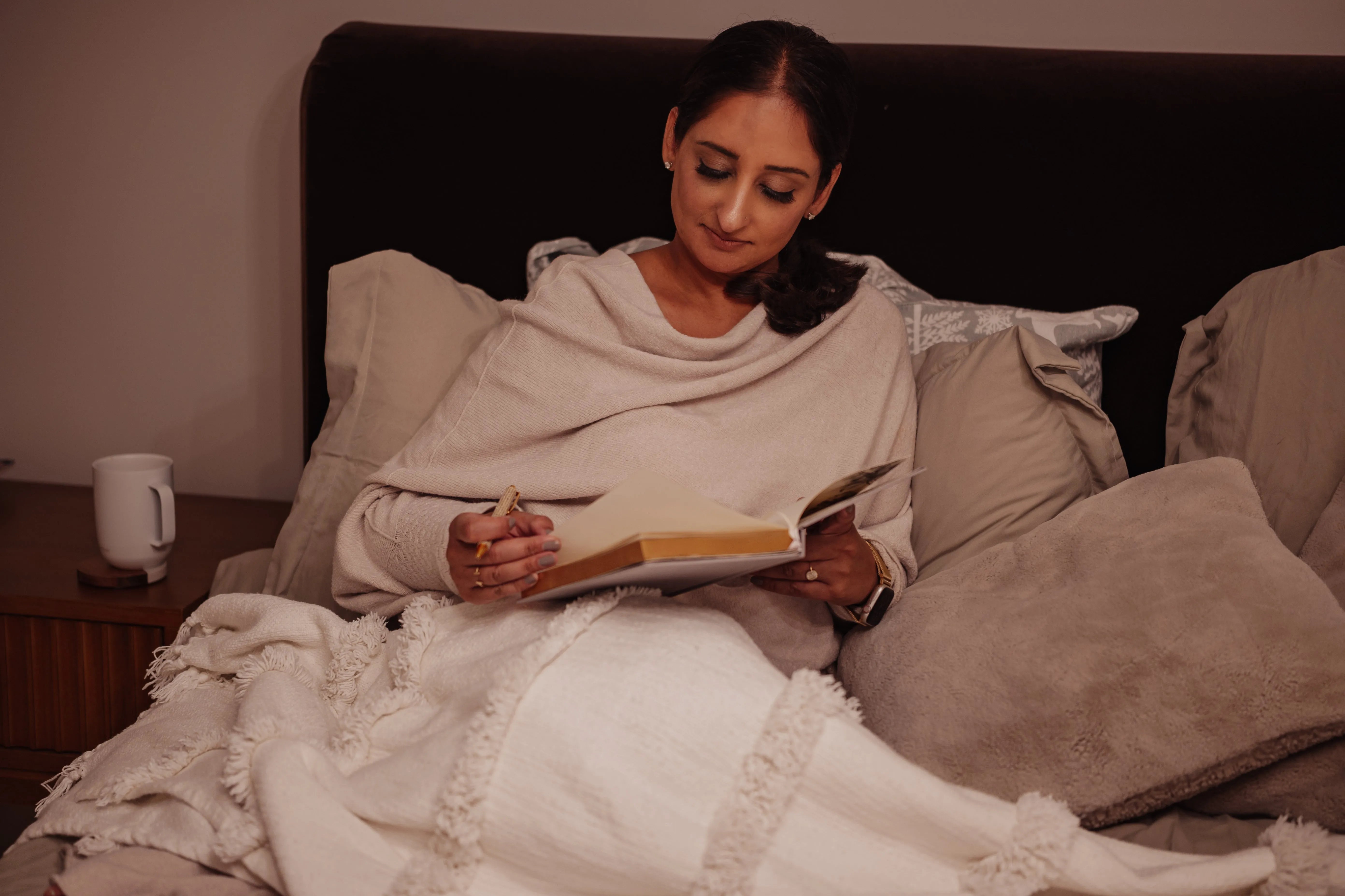 Woman reading a book on a bed with a warm blanket and soft lighting.