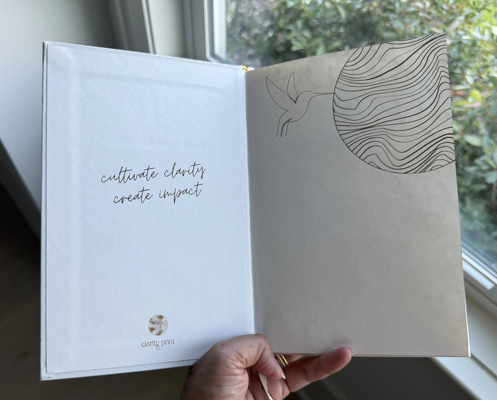 Hand holding the .1 Daily Gratitude Journal with gold foil design and slogan, against a window background